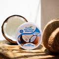 A round white container of Fiabina Nail Polish Remover in Coconut Scents, styled on a wooden surface with a halved coconut on the left and a whole coconut on the right.