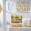 Fiabila Acne & Freckle Soap box displayed on a bathroom counter with a shower head, tiled wall, and folded towels in the background, with large promotional text reading 'ACNE & FRECKLE SOAP'.